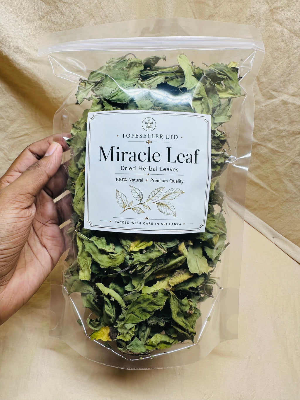 A hand holds a clear plastic bag filled with dried green herbal leaves. The front label reads 'TOPESELLER LTD Miracle Leaf Dried Herbal Leaves' on a beige fabric background.